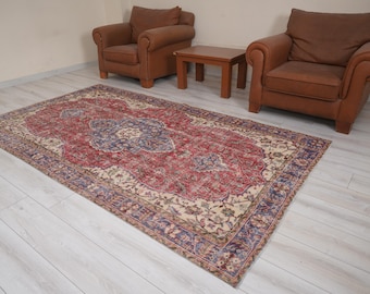 May include: A rectangular, vintage-style rug with a central medallion design in shades of red, blue, and cream. The rug features an ornate border and is placed on a light-colored wooden floor. Two brown armchairs and a small wooden table are in the background.