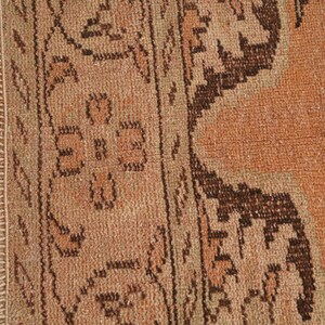 May include: Close-up of a section of a vintage rug with a distressed, faded appearance. The rug features a warm color palette of orange, brown, and beige, with intricate geometric and floral patterns. The rug's edge has a fringe.