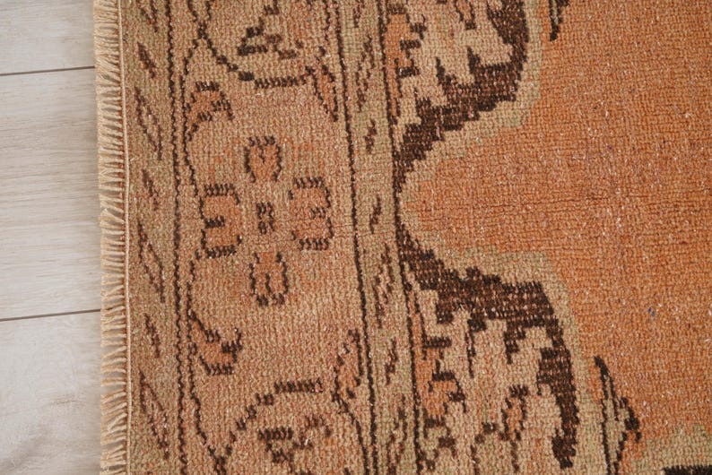 May include: Close-up of a section of a vintage rug with a distressed, faded appearance. The rug features a warm color palette of orange, brown, and beige, with intricate geometric and floral patterns. The rug's edge has a fringe.