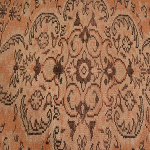 May include: Close-up of a vintage rug with an intricate, symmetrical pattern. The rug features a warm, peachy-orange background with dark brown floral and geometric designs. The detailed pattern creates a rich, textured appearance.