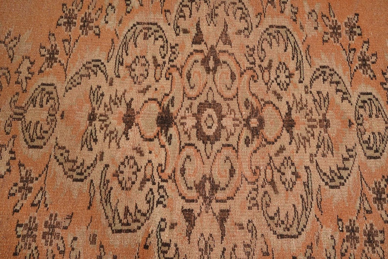 May include: Close-up of a vintage rug with an intricate, symmetrical pattern. The rug features a warm, peachy-orange background with dark brown floral and geometric designs. The detailed pattern creates a rich, textured appearance.