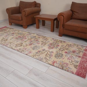 May include: A rectangular, floral patterned rug with a cream-colored background and red border. The rug features a faded floral design in shades of pink, blue, and green. Two brown armchairs and a small wooden table are in the background.