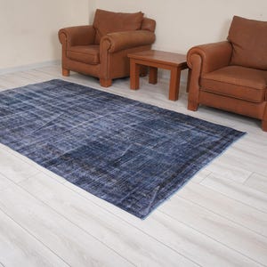 Pode incluir: Um tapete vintage azul e cinza desbotado com um visual desbotado. O tapete está colocado sobre um piso de madeira branca com duas poltronas de couro marrom e uma mesa de centro de madeira.