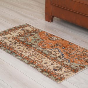 May include: A rectangular area rug featuring a distressed pattern in shades of burnt orange, cream, sage green, and navy blue. The design includes a central field with floral motifs surrounded by a wide, intricate border with geometric patterns.