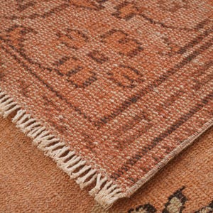 May include: Close-up of a section of a woven rug with a geometric pattern. The rug features a warm color palette of orange and brown tones. The edges are finished with a fringe.