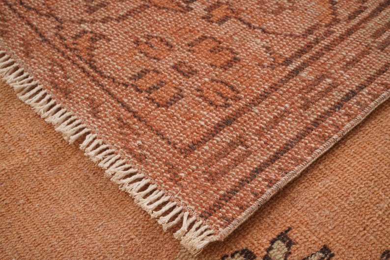 May include: Close-up of a section of a woven rug with a geometric pattern. The rug features a warm color palette of orange and brown tones. The edges are finished with a fringe.