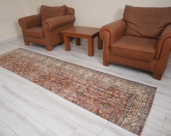 May include: A long, rectangular rug with a distressed, vintage look. The rug features a complex floral pattern in shades of red, brown, and cream. Two brown leather armchairs and a small wooden table are in the background.
