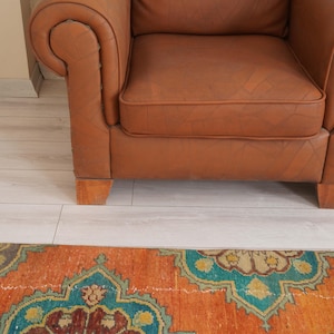 May include: A brown leather armchair with a tufted seat and rolled arms. An orange and teal patterned rug is in the foreground. The chair is on wooden legs.