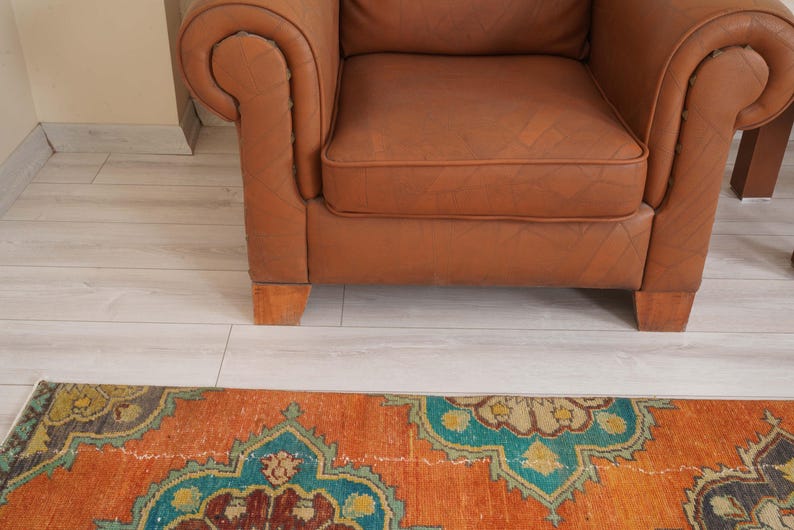 May include: A brown leather armchair with a tufted seat and rolled arms. An orange and teal patterned rug is in the foreground. The chair is on wooden legs.