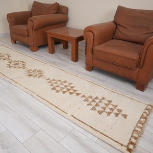 May include: A long, rectangular rug with a cream-colored background and geometric brown patterns. The rug is placed on a light-colored wooden floor, with two brown leather armchairs and a small wooden table in the background.