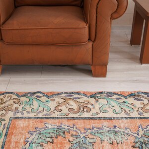 Pode incluir: Um tapete vintage com um padr&atilde;o floral em tons de azul, verde e laranja. O tapete tem uma borda de cor creme e est&aacute; colocado sobre um piso de madeira clara.