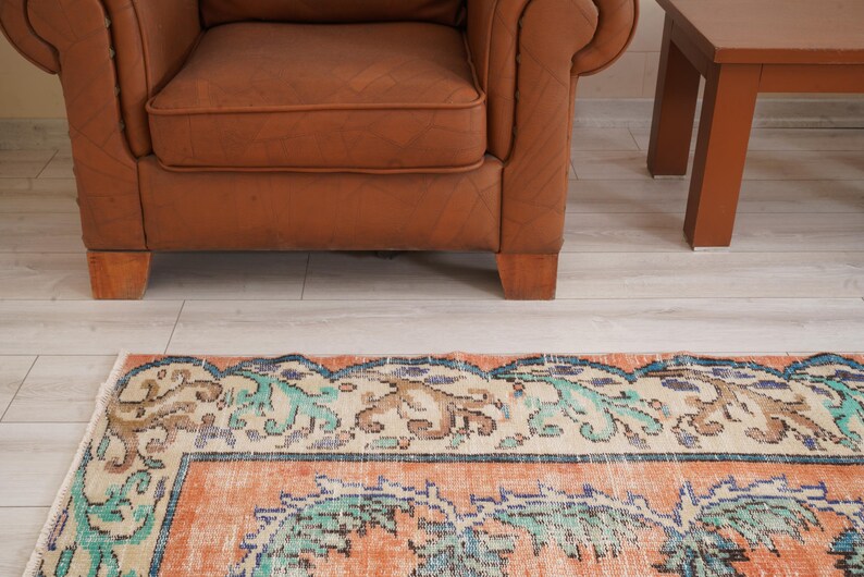 Pode incluir: Um tapete vintage com um padr&atilde;o floral em tons de azul, verde e laranja. O tapete tem uma borda de cor creme e est&aacute; colocado sobre um piso de madeira clara.