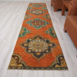 May include: An orange runner rug with a detailed, symmetrical pattern in teal, navy, and gold. The rug is rectangular and appears to be made of woven material. The design features floral and geometric elements, creating a vintage aesthetic. The rug measures approximately 3 feet wide.