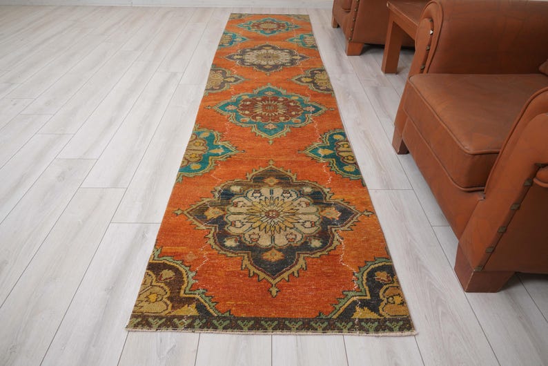 May include: An orange runner rug with a detailed, symmetrical pattern in teal, navy, and gold. The rug is rectangular and appears to be made of woven material. The design features floral and geometric elements, creating a vintage aesthetic. The rug measures approximately 3 feet wide.