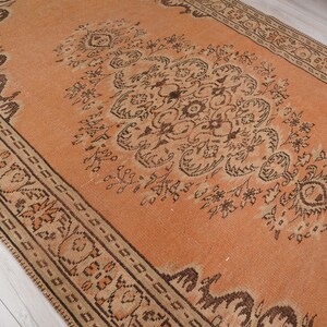 May include: An orange and brown vintage rug with an intricate floral pattern. The rug has a central design with a border, and is likely made of wool or a similar material. The rug is rectangular and appears to be in good condition.