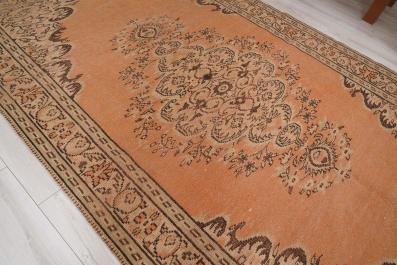 May include: An orange and brown vintage rug with an intricate floral pattern. The rug has a central design with a border, and is likely made of wool or a similar material. The rug is rectangular and appears to be in good condition.
