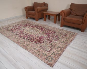 May include: A rectangular, vintage-style rug with a central maroon field and ornate floral patterns in beige, green, and cream. The rug is placed on a light-colored wooden floor, with two brown leather armchairs and a small wooden table in the background.