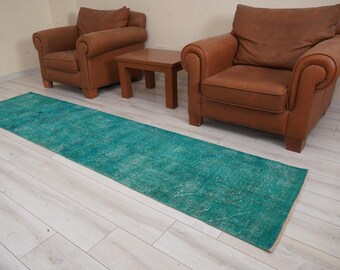 May include: A teal blue runner rug with a distressed, vintage look. The rug is laid out on a light wood floor between two brown leather armchairs.