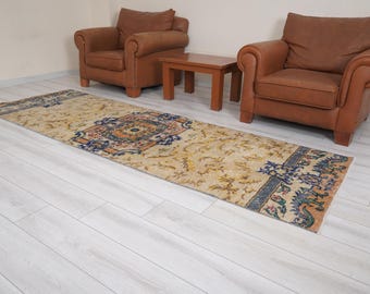 May include: A beige and cream runner rug with a floral pattern and blue accents. The rug is laid out on a white wood floor in front of two brown leather armchairs and a wooden coffee table.
