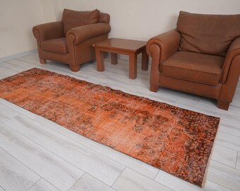 May include: An orange and brown distressed runner rug with a floral pattern. The rug is placed on a light wood floor, with two brown leather armchairs and a small wooden table in the background. The rug's design is faded.