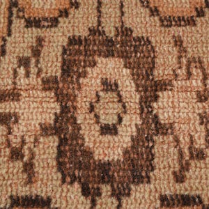 May include: Close-up of a woven rug with a repeating geometric pattern. The rug features a warm color palette of brown, tan, and peach tones. The design includes stylized floral or medallion shapes.