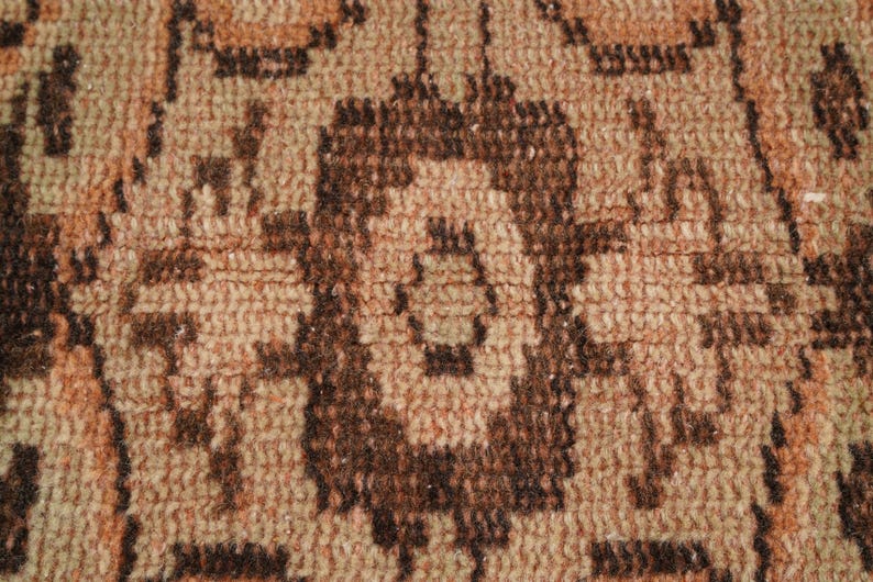 May include: Close-up of a woven rug with a repeating geometric pattern. The rug features a warm color palette of brown, tan, and peach tones. The design includes stylized floral or medallion shapes.