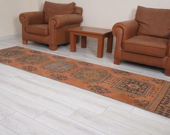 Op de afbeelding: Een lang, rechthoekig, oranje en bruin patroonkleed met een geometrisch ontwerp. Het kleed ligt op een witte houten vloer tussen twee bruine leren fauteuils en een houten salontafel.