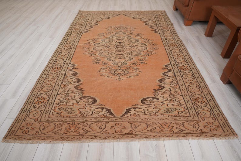 May include: A rectangular, vintage-style rug with an intricate floral pattern in shades of peach and brown. The rug features a central medallion design and a detailed border. The rug is placed on a light-colored wooden floor, showcasing its warm tones and traditional aesthetic.