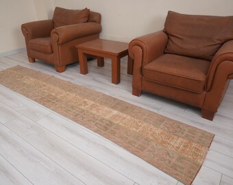 May include: A long, rectangular rug with a faded, patterned design in shades of beige, brown, and green. Two brown leather armchairs and a small wooden table are in the background, set against a light-colored wall and floor.