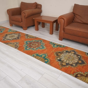 May include: An orange runner rug with a repeating floral pattern in teal, brown, and gold. The rug is rectangular and placed on a light wood floor, with two brown leather armchairs and a small wooden table in the background.