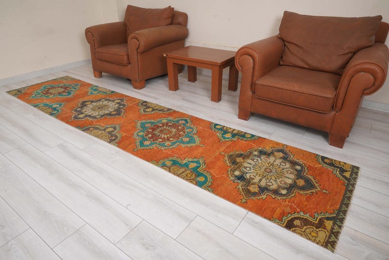 May include: An orange runner rug with a repeating floral pattern in teal, brown, and gold. The rug is rectangular and placed on a light wood floor, with two brown leather armchairs and a small wooden table in the background.
