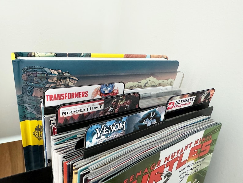 Peut inclure: Une pile de bandes dessin&eacute;es avec des s&eacute;parateurs &eacute;tiquet&eacute;s "Transformers", "Blood Hunt", "Venom" et "Ultimate Spider-Man". La bande dessin&eacute;e du dessus a une reliure jaune et le texte "GIS" sur la couverture.