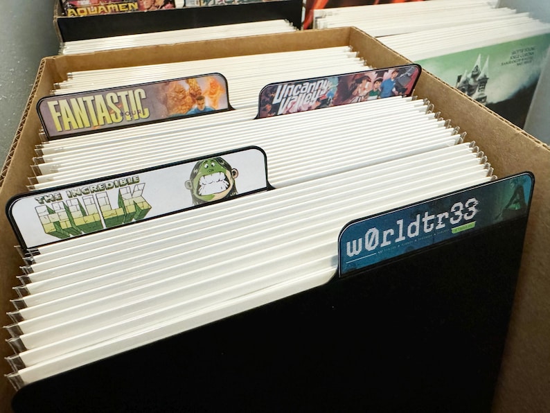 Peut inclure: Une bo&icirc;te de rangement noire avec des s&eacute;parateurs et des &eacute;tiquettes blancs. Les &eacute;tiquettes comprennent "Fantastic", "The Incredible Hulk", "Uncanny X-Men", et "Worldtr33".