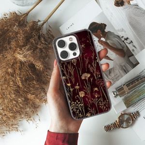 May include: A smartphone case featuring a burgundy backdrop with a gold floral pattern. The case has a transparent edge and a camera aperture. The case is held by a person.