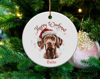 May include: A round white ceramic Christmas ornament featuring a brown dog wearing a red and white Santa hat. The text Merry Woofmas is printed in brown above the dog, and the name Bella is printed in red below the dog, surrounded by snowflakes.