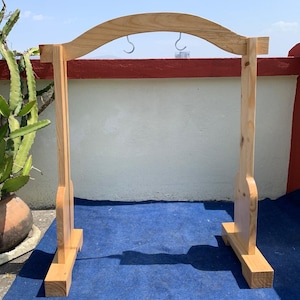 May include: A wooden display stand with a curved top and two metal hooks. The stand is made of light-colored wood and has a rectangular base. The background includes a potted cactus and a blue surface.