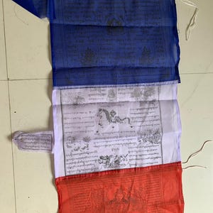 May include: A traditional Tibetan prayer flag with three panels. The top panel is blue with black text and images. The middle panel is white with black text and images. The bottom panel is red with black text and images. The flag is made of thin fabric and has a string at the top for hanging.