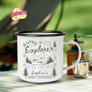 May include: White enamel camping mug with a black rim and handle. The mug features a green and white graphic with the text "A Little Explorer is on the Way Sophia's Baby Shower". The design includes mountains, trees, and a compass.