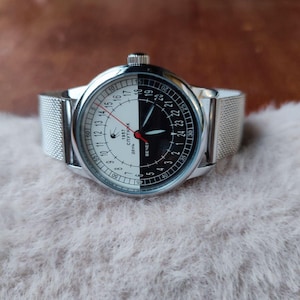 Puede incluir: Un reloj de pulsera plateado con una esfera única en blanco y negro. La esfera del reloj está dividida por la mitad, con un lado blanco y el otro negro. El reloj tiene una correa de malla y un segundero rojo. El texto "1957 Sputnik" es visible.