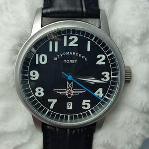 May include: A black-faced wristwatch with white numerals and hands, and a silver-colored case. The watch face includes the text "Штурманские ПОЛЕТ" and a red star emblem. The watch has a black leather band.
