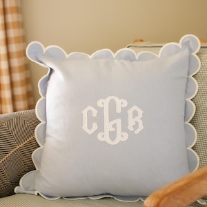 Pode incluir: Uma almofada quadrada azul-clara com uma borda recortada branca e um monograma branco bordado no centro, apresentando as letras C, G e B entrelaçadas num estilo de fonte cursiva decorativa na parte frontal da capa de tecido.