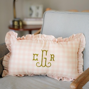 May include: Pink gingham cushion with frilled edges and gold embroidered initials. The rectangular cushion is on a grey and white checked chair. A decorative accent for a bedroom or living room.