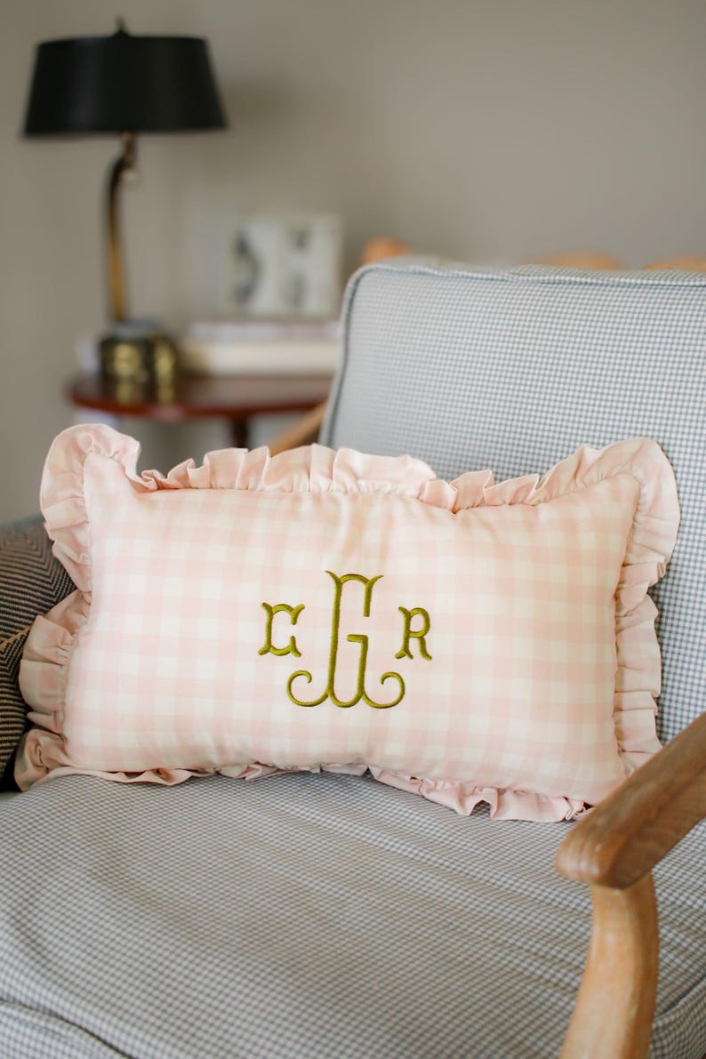 May include: Pink gingham cushion with frilled edges and gold embroidered initials. The rectangular cushion is on a grey and white checked chair. A decorative accent for a bedroom or living room.