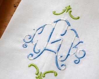 May include: White linen napkin with embroidered monogram in blue and white stripes, and green accents. The monogram features elegant, swirling letters, and is framed by decorative green flourishes. The napkin has a delicate, stitched border.