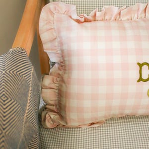 May include: A pink and white gingham cushion with a frilled edge sits on a grey and white checked chair. The cushion has gold embroidered lettering. The chair has a wooden frame and a blue and white patterned fabric.