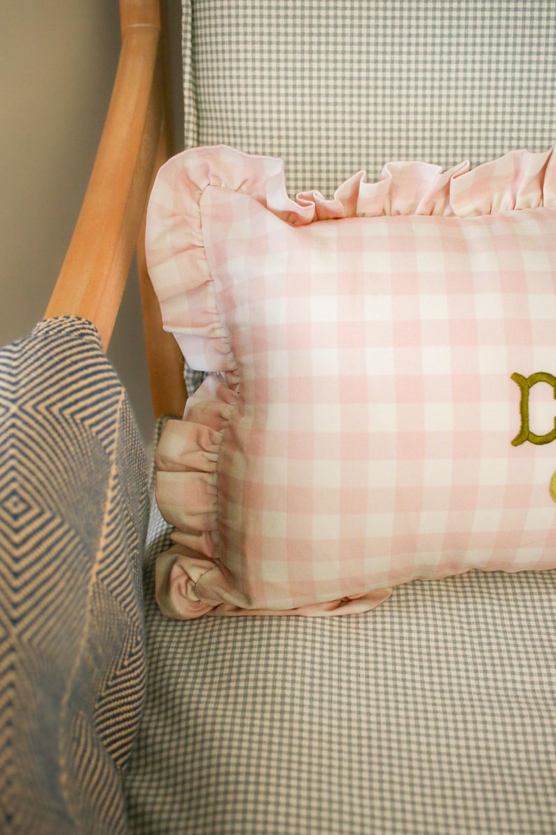 May include: A pink and white gingham cushion with a frilled edge sits on a grey and white checked chair. The cushion has gold embroidered lettering. The chair has a wooden frame and a blue and white patterned fabric.