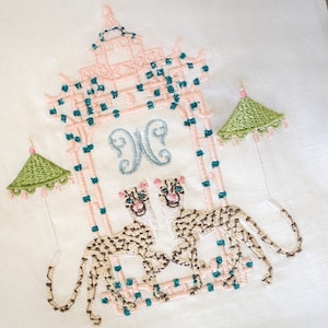 Op de afbeelding: Een geborduurde witte linnen theedoek met een roze pagode-ontwerp, twee groene paraplu's en twee bruine en witte luipaarden. De pagode heeft een blauw en wit monogram "N" in het midden.