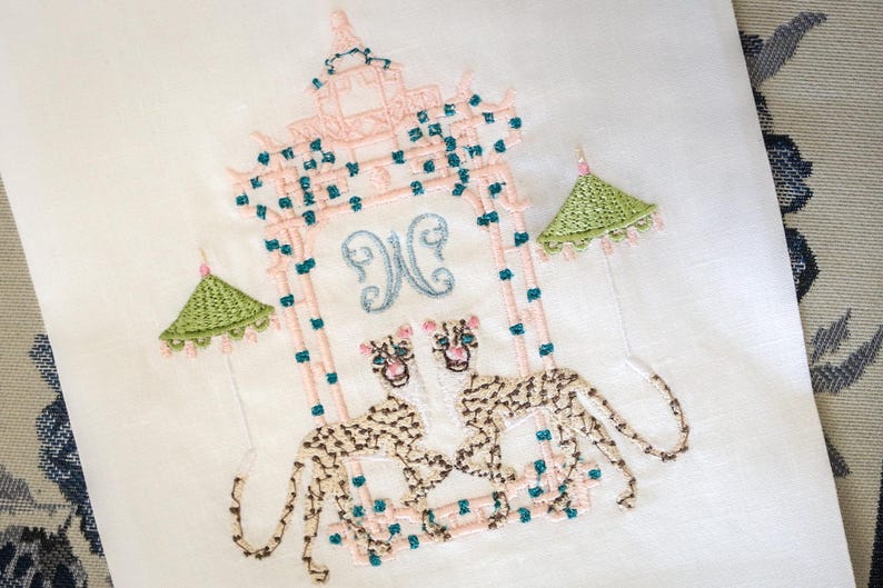 Op de afbeelding: Een geborduurde witte linnen theedoek met een roze pagode-ontwerp, twee groene paraplu's en twee bruine en witte luipaarden. De pagode heeft een blauw en wit monogram "N" in het midden.