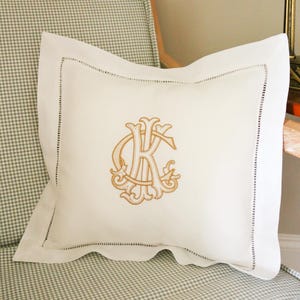Puede incluir: Un cojín decorativo blanco con un monograma bordado en oro, con la letra "K" en un diseño ornamentado. El cojín tiene un borde cosido y está colocado sobre una silla a cuadros.