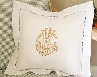 May include: A white cushion with a gold embroidered monogram, featuring the letter "K" in an ornate design. The cushion has a stitched border and is placed on a checkered chair.